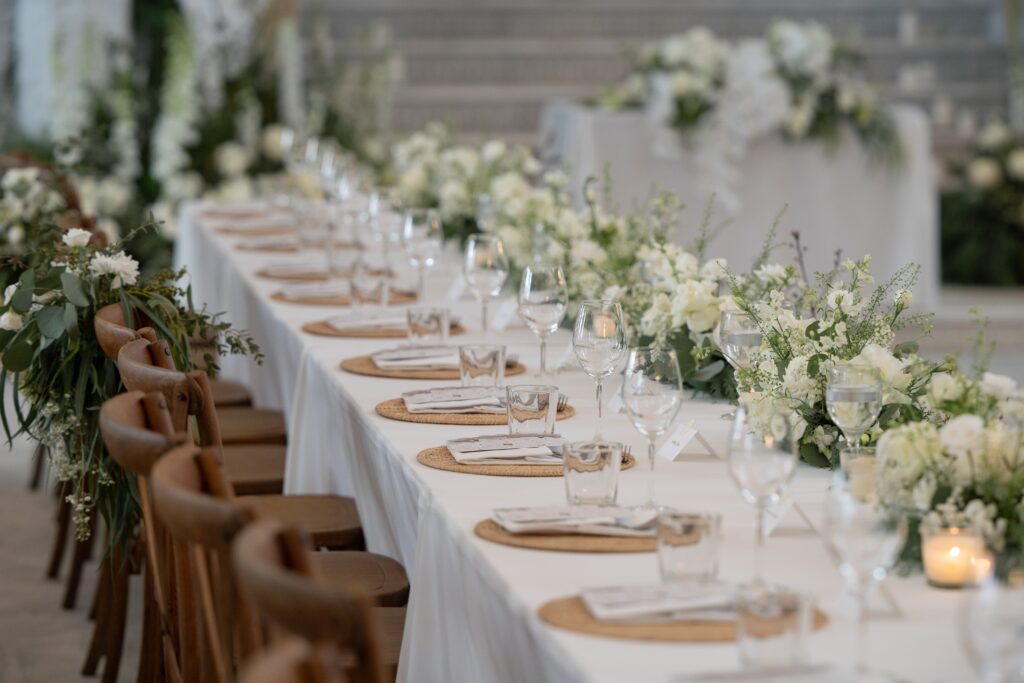Lange Hochzeitstafel mit weißen Blumen, Eukalyptus und Kerzen, davor Holzstühle
