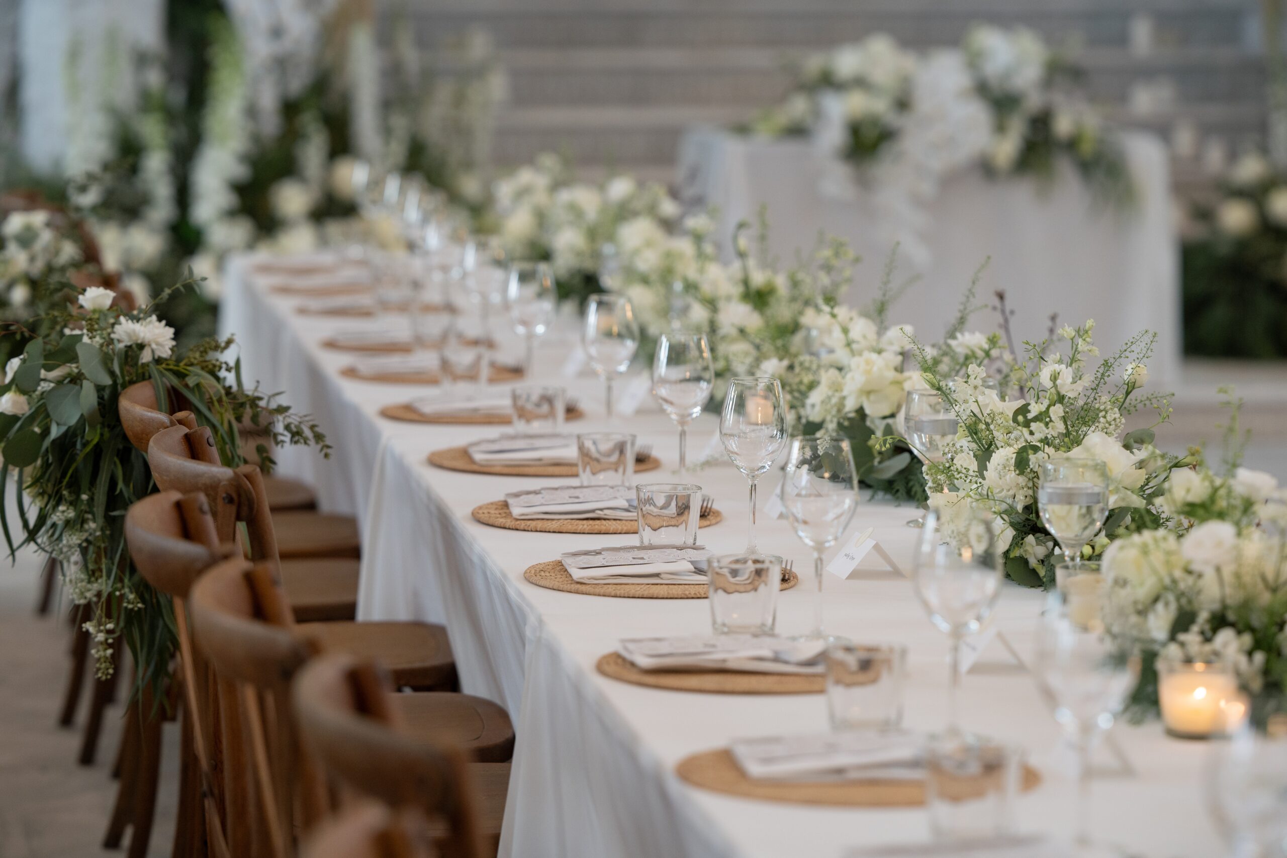 Lange Hochzeitstafel mit weißen Blumen, Eukalyptus und Kerzen, davor Holzstühle