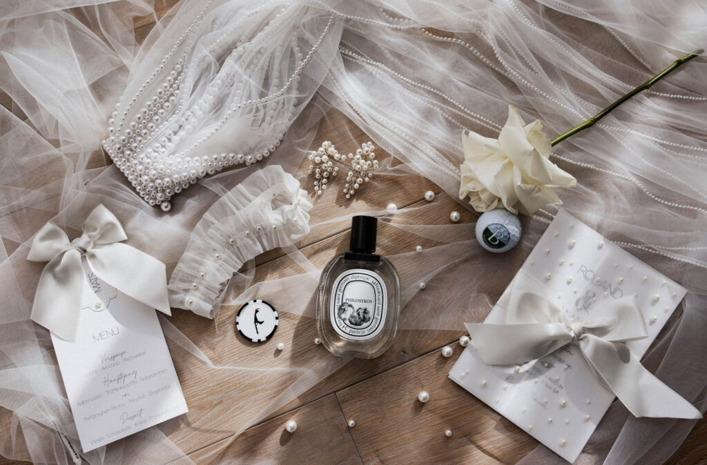 Flatlay mit Brautschleier, Perlenschmuck, Strumpfband, Parfum, Menükarte und weißer Rose als Details einer Hochzeit auf Sylt