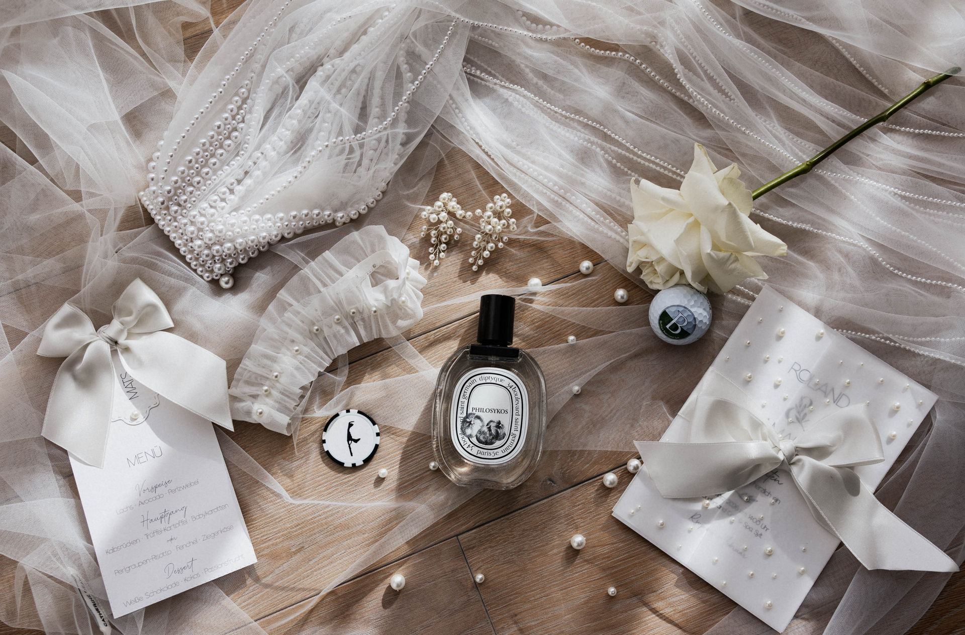 Flatlay mit Brautschleier, Perlenschmuck, Strumpfband, Parfum, Menükarte und weißer Rose als Details einer Hochzeit auf Sylt
