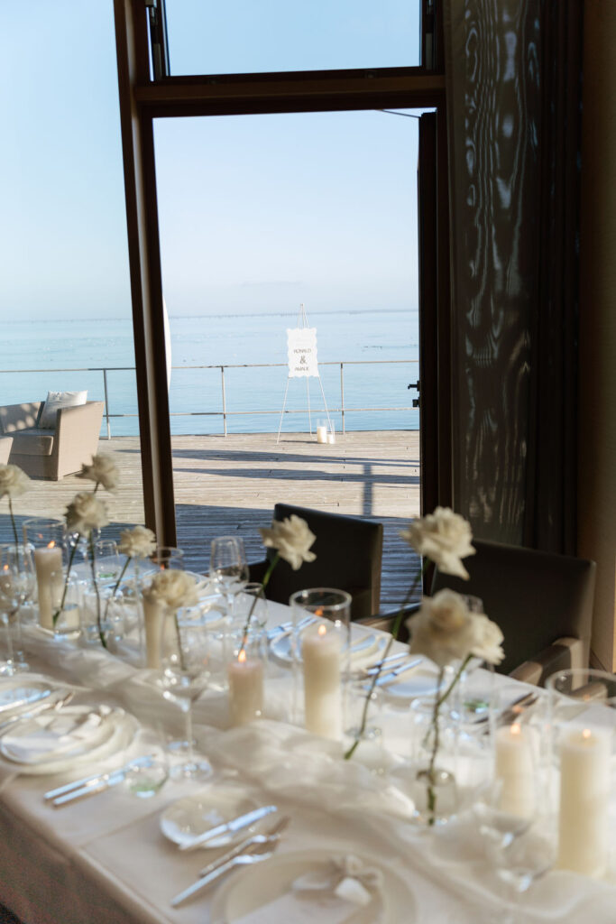 Hochzeitstafel mit weißen Rosen und Blick durch die Fensterfront auf das Meer