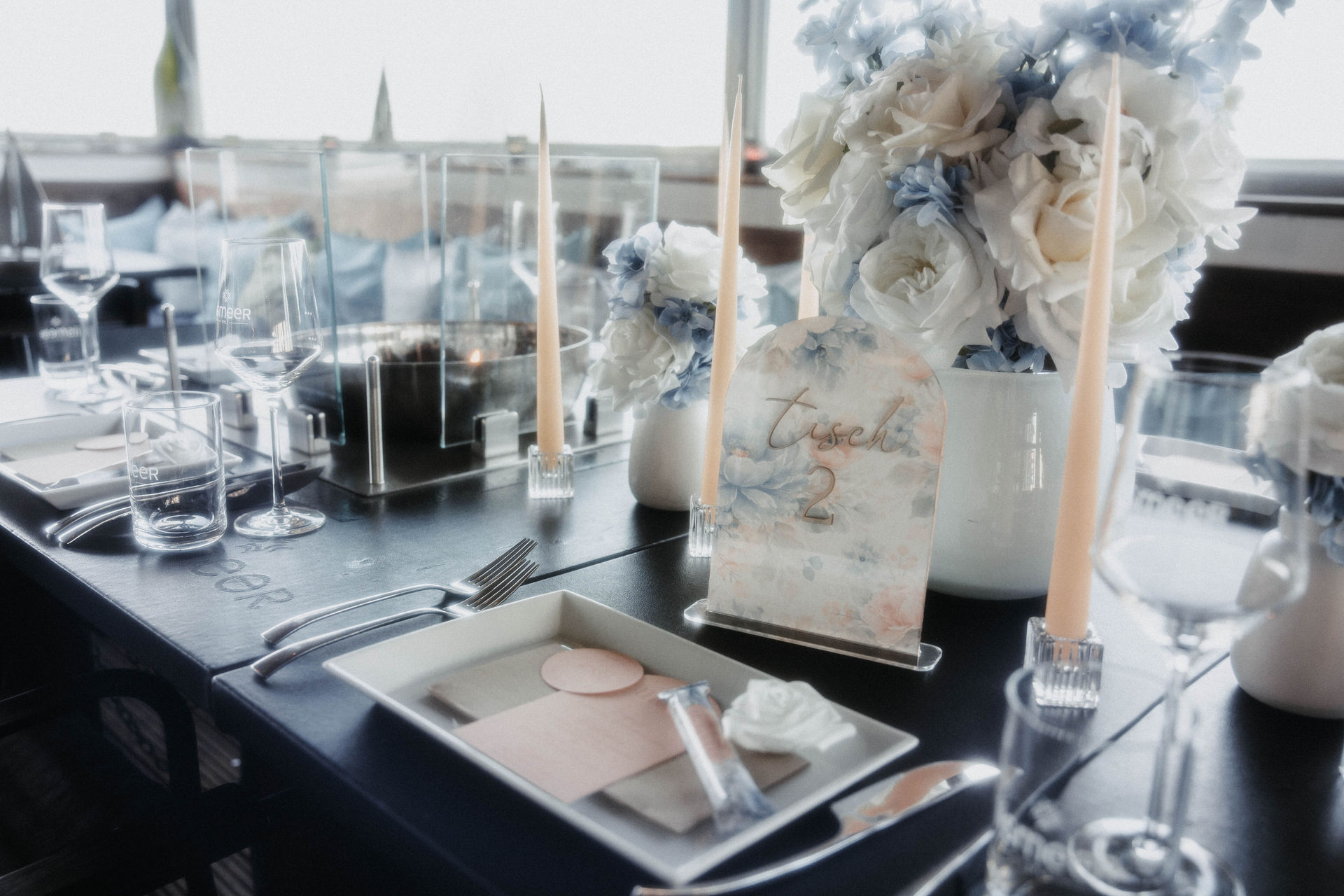 Gedeckter Hochzeitstisch mit handgemalter Tischkarte, weißen Rosen und hellblauen Blüten