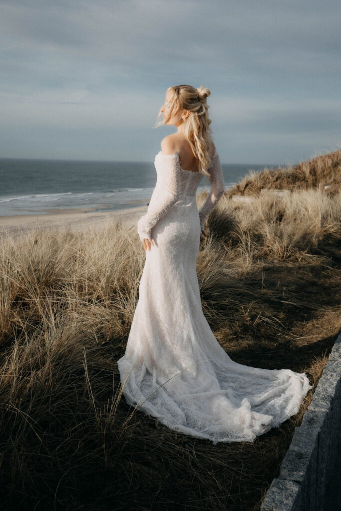 Braut im Spitzenkleid steht in den Dünen und blickt auf das Meer