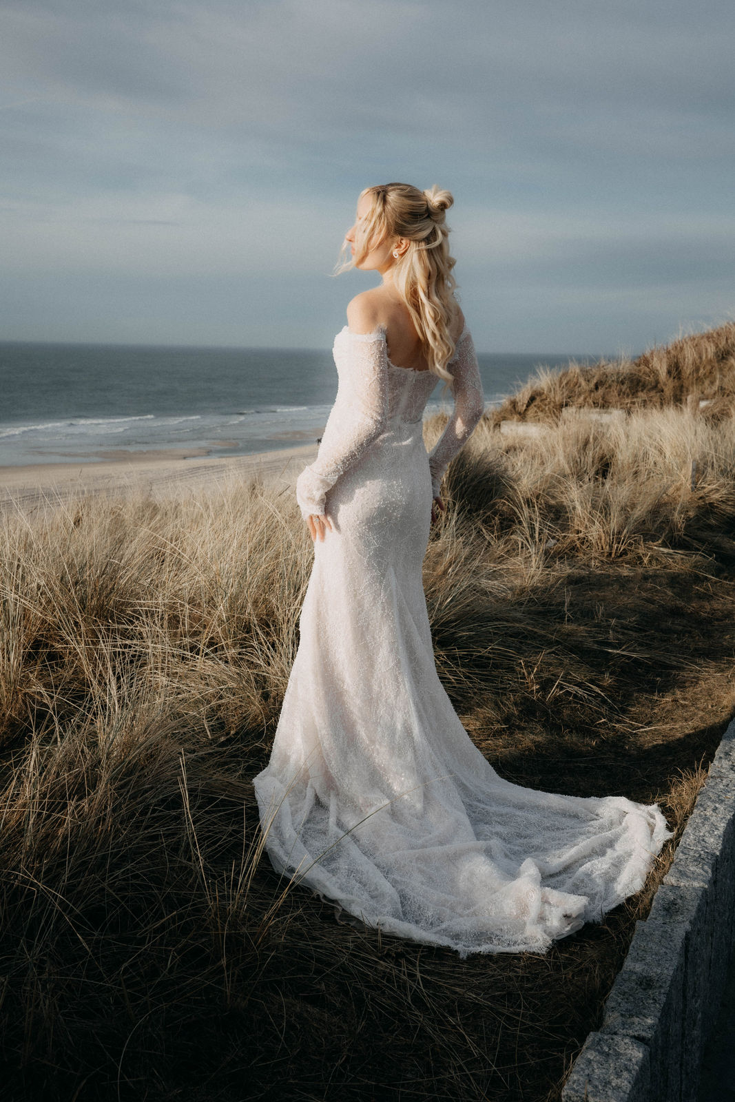 Braut im Spitzenkleid steht in den Dünen und blickt auf das Meer