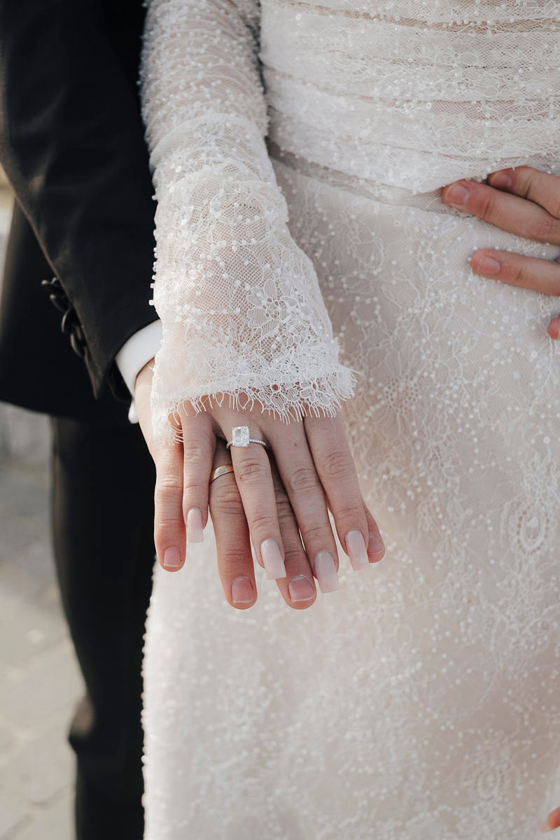 Hände des Brautpaars auf dem Spitzenkleid, im Detail der Verlobungsring