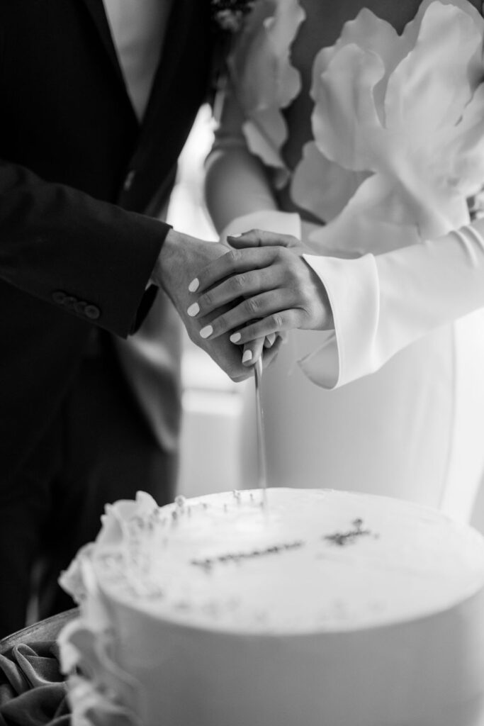 Hände des Brautpaars beim Anschneiden der weißen Hochzeitstorte